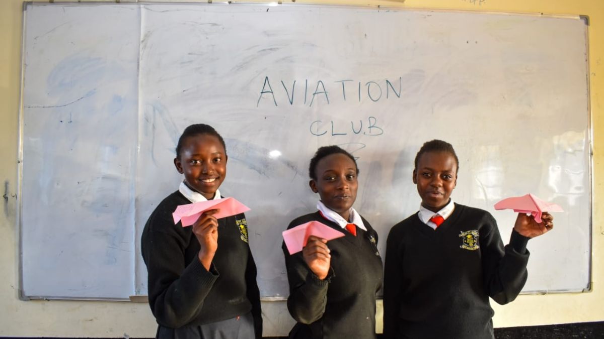 Three Kenyan girls in school uniforms in front of a white board that says "Aviation Club", holding pink paper airplanes