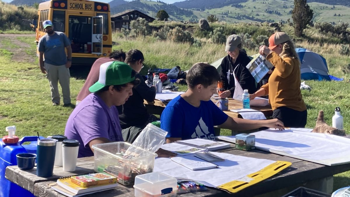 Students outside on a picnic table writing on flip charts, with a school bus and tents in the background
