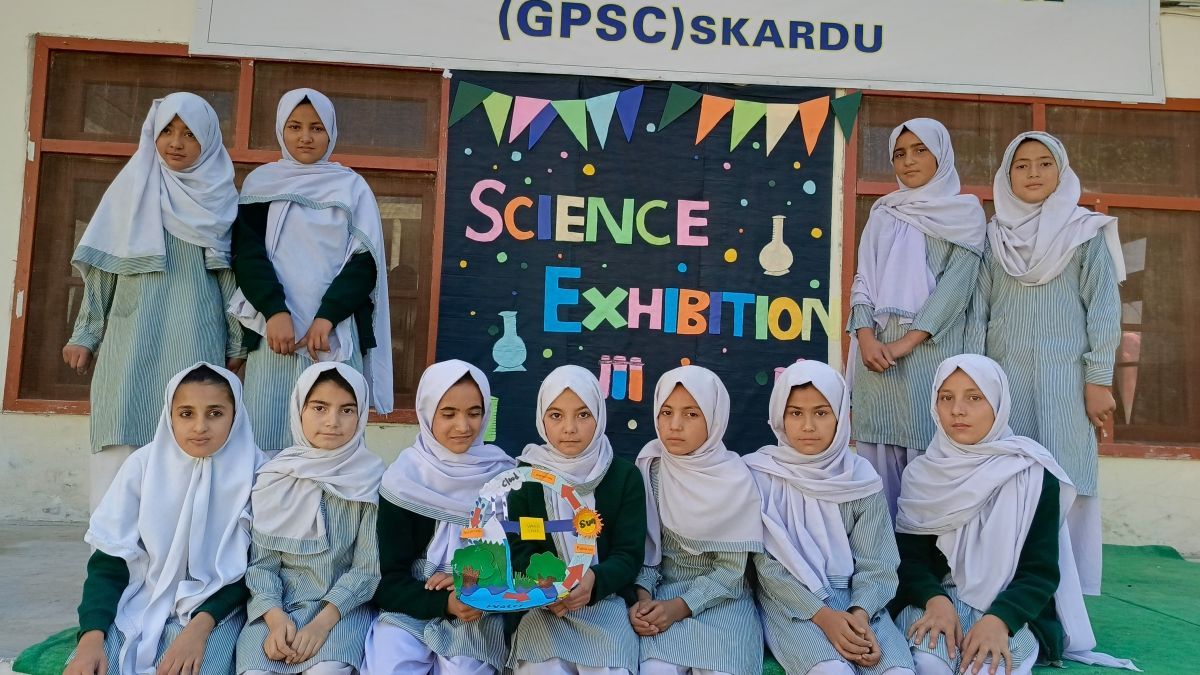 A group of Pakistani school girls posing in front of a colorful poster that says "Science Exhibition", with two students holding a water cycle diorama