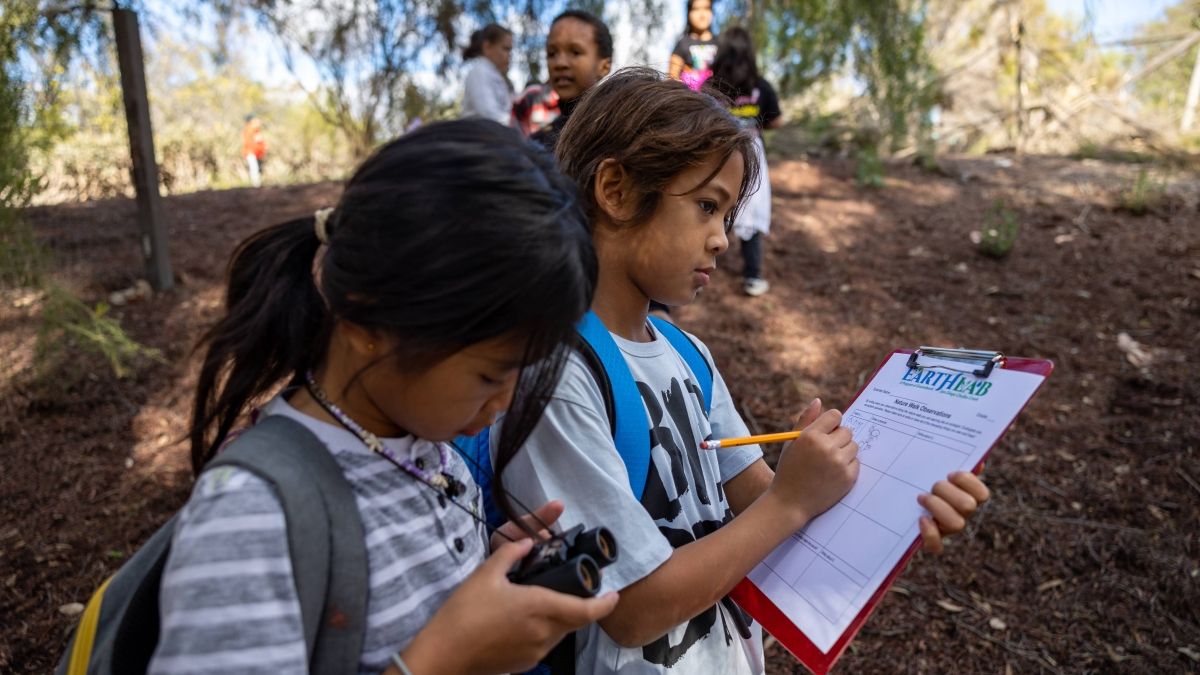 Two young students outside, one holding binoculars and one writing on a clipboard that says Earth Lab