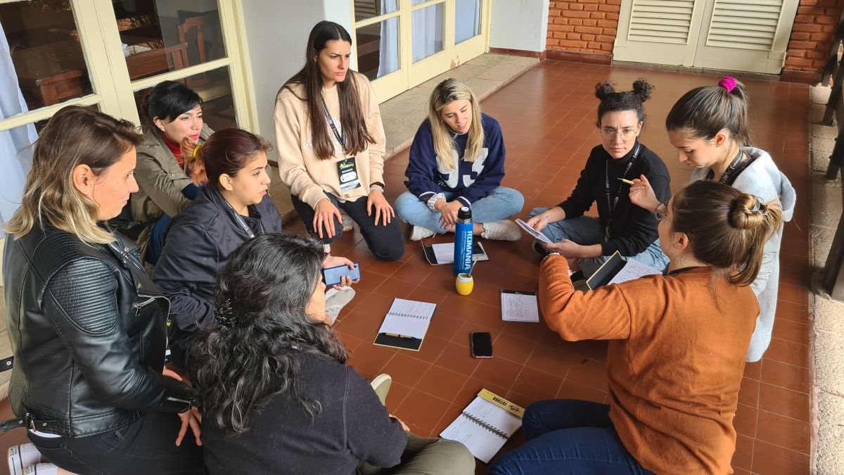 A group of young women sitting in a circle on a patio, talking and taking notes.