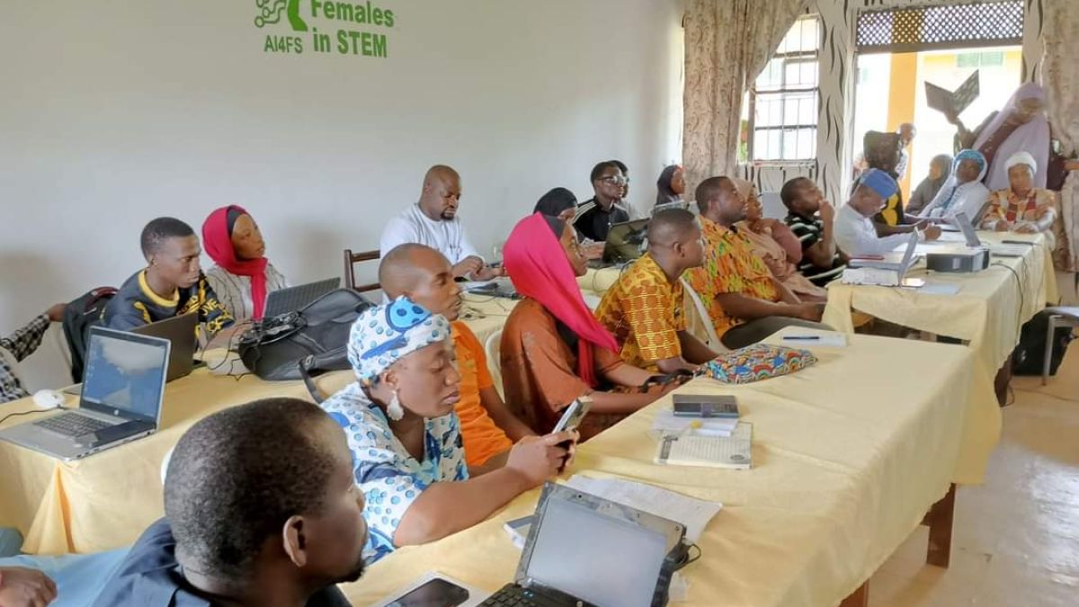 A large group of Nigerian adults sitting along tables watching a presentation, with "AI for Females in STEM" on the wall behind them