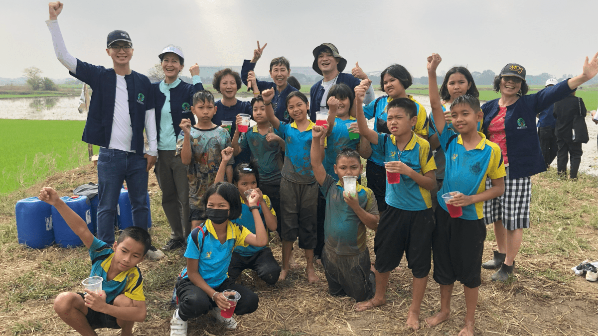 A group of children cheering, posing for a photo outside next to a rice field