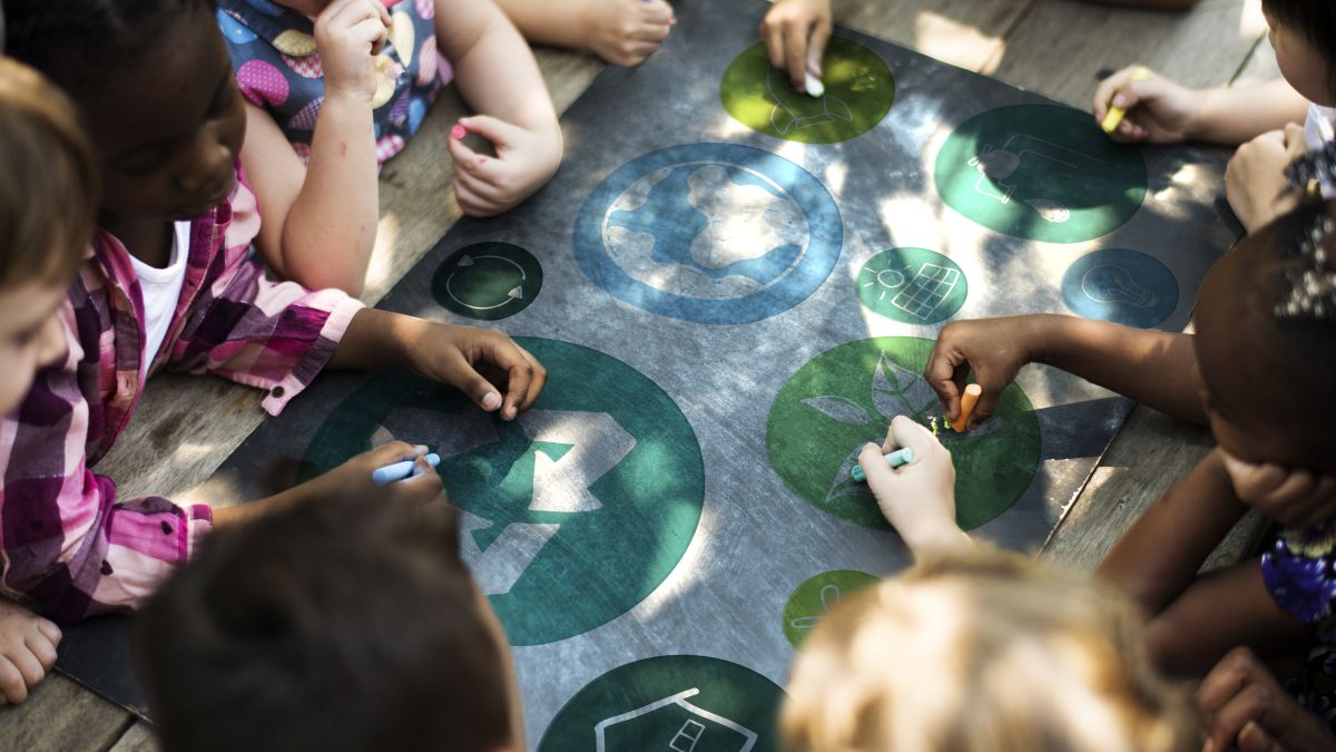 Young students holding chalk sit around a table with sustainability symbols drawn on it.
