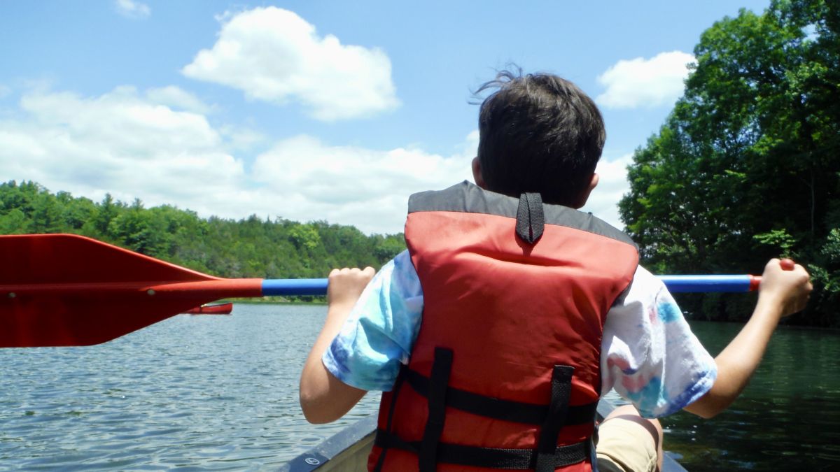 A kid wearing a red life vest paddles on a forest-lined body of water