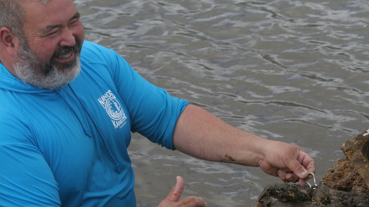 A man stands in water and gestures toward a clip on a rock