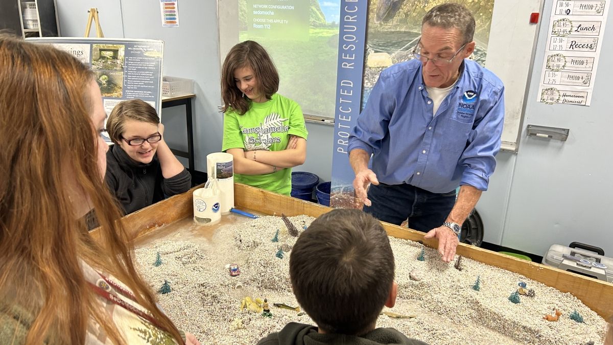 Group of students gather around a NOAA educator to learn about a water table