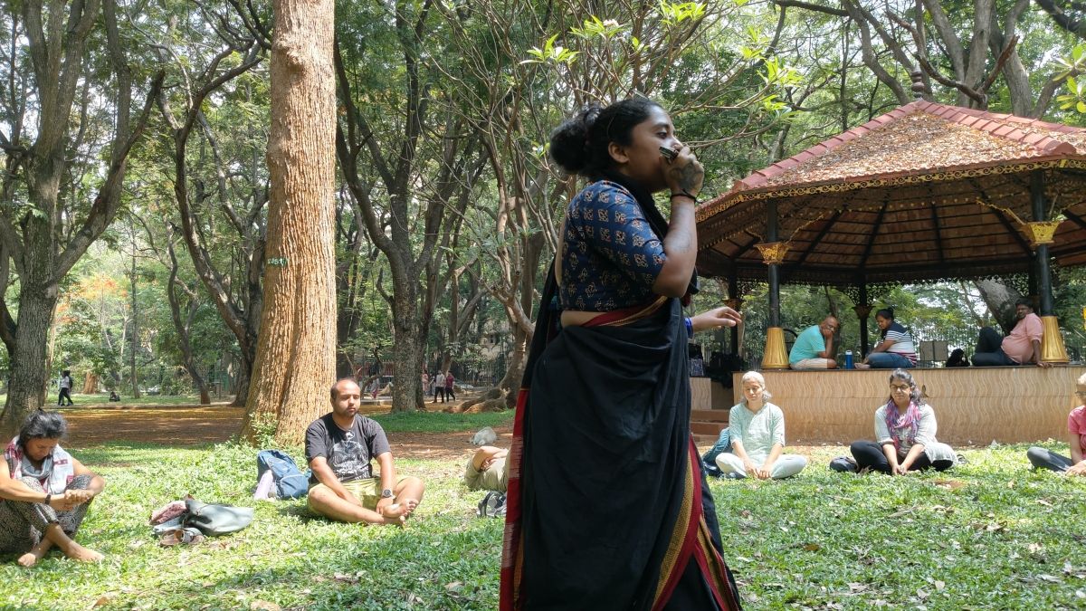 A woman standing in the center of a half-circle of people sitting on the grass with their eyes closed