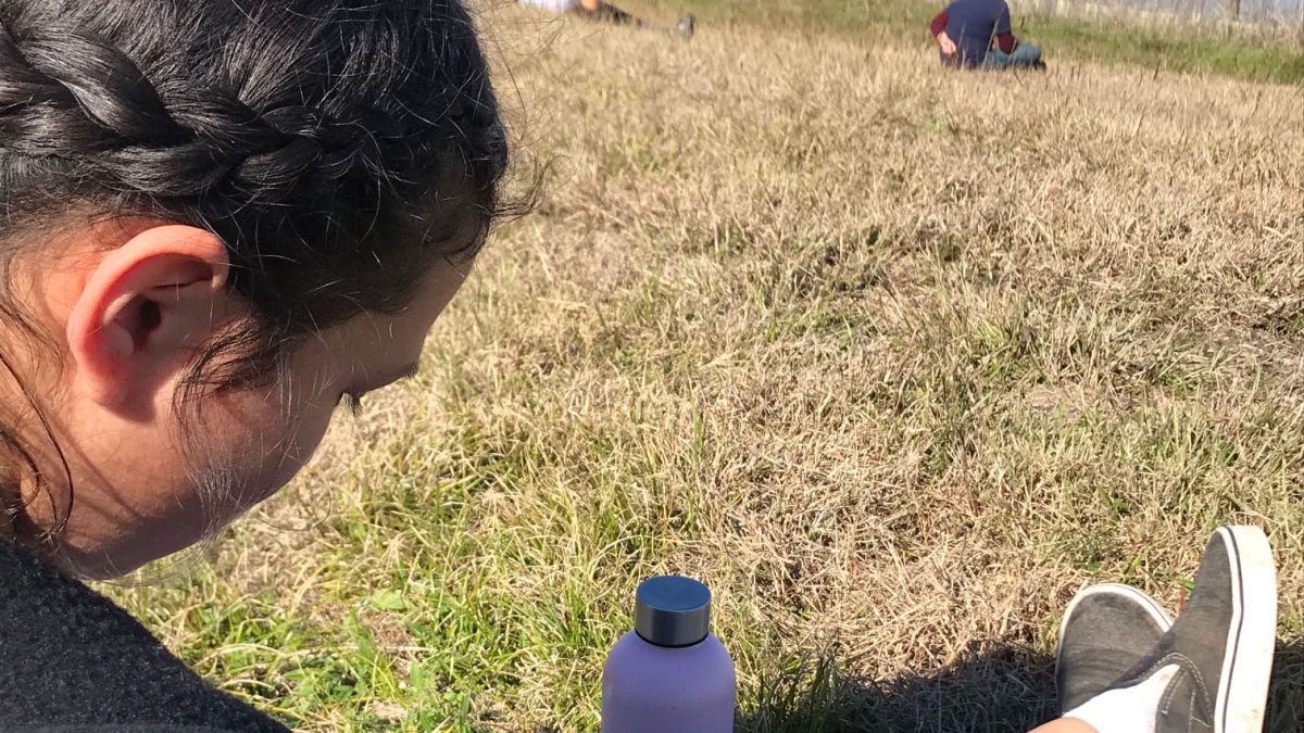 girl sits on grass by water writing in notebook