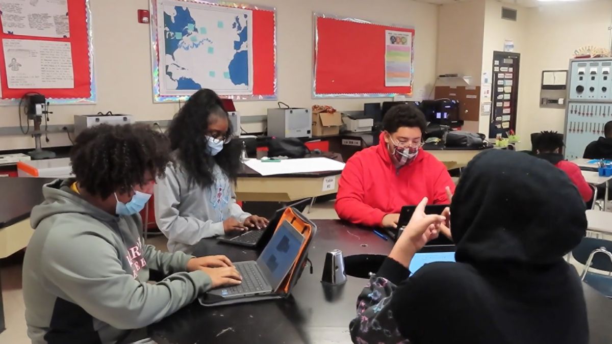 four students wearing masks working on computers in a group in a classroom