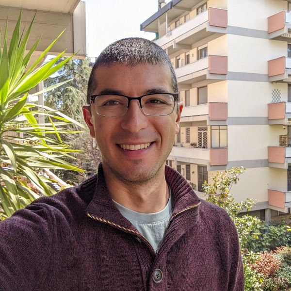 A person outside wearing glasses and a burgundy sweater with a tall building in the background