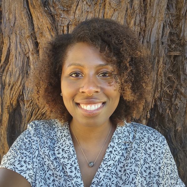 A person wearing a white blouse with a black leaf pattern smiling in front of a tree