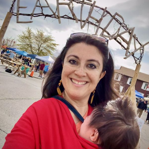 A closeup of Alma Alicia Padilla holding a baby in a red wrap while at an outdoor Earth Day event
