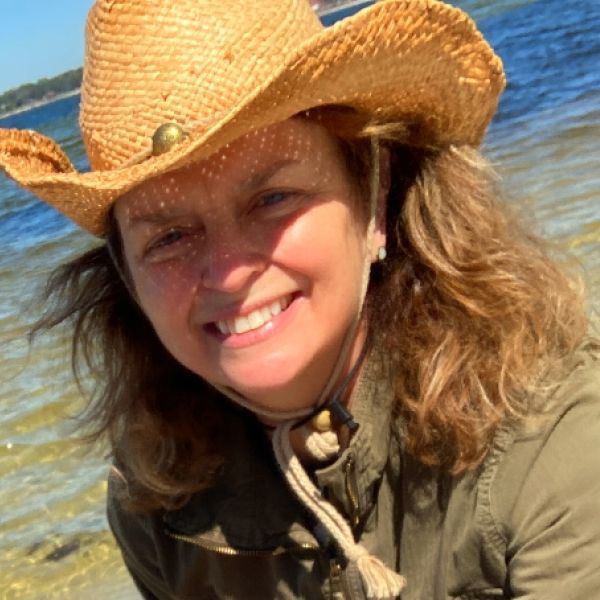 A woman with shoulder-length hair, wearing a cowboy hat, sits in front of water
