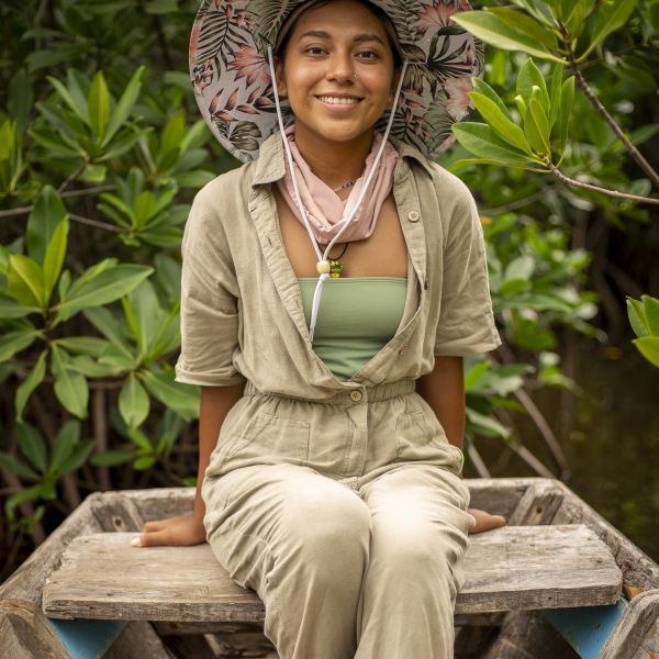 EE 30 Under 30 Awardee Jabel Gomez sits on a wooden bench while wearing a sun hat and sandals, green plants behind her