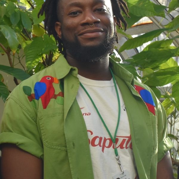 EE 30 Under 30 Awardee Jibreel Cooper wearing an open green button-up with parrots on the shoulders, standing in front a green leafy plant