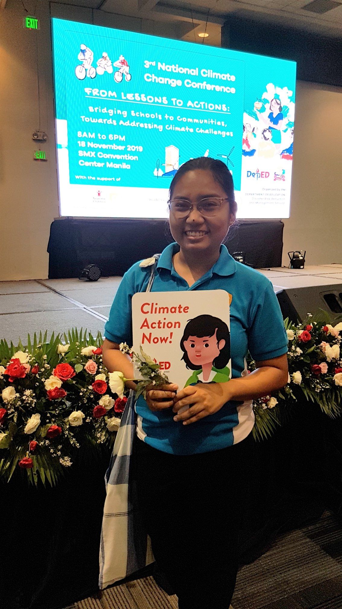A picture of a smiling woman posing in front of a stage and holding a book that says "Climate Action Now"