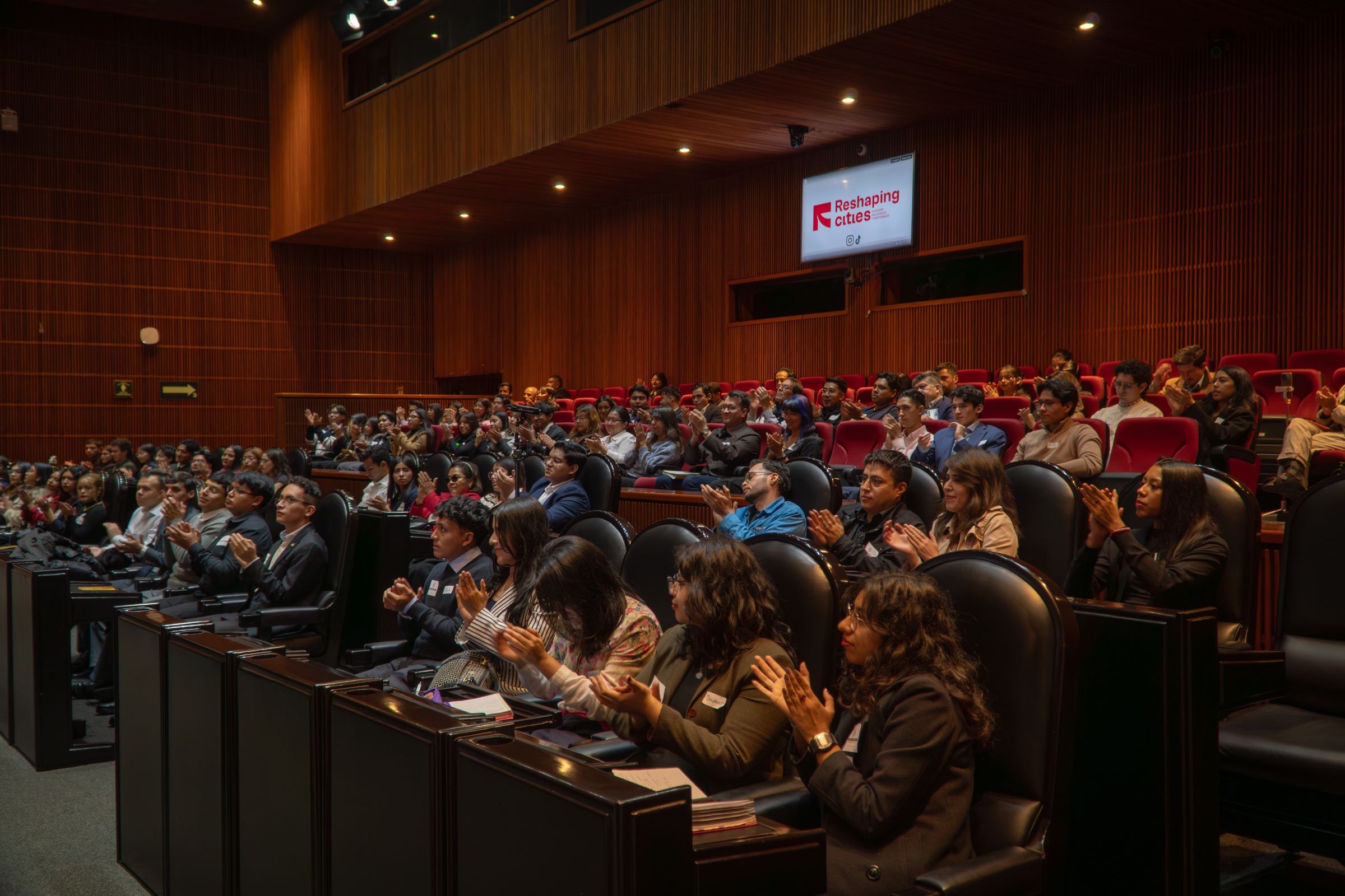 An audience of university students inside a theater listening to a presentation.