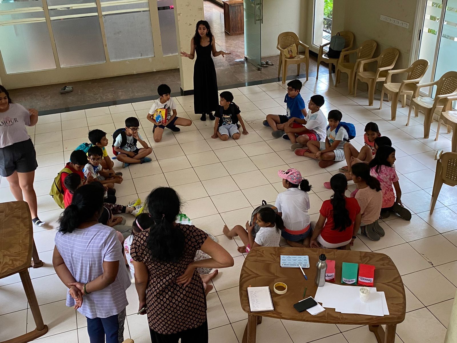 A group of children in a circle with a woman standing and speaking to them.