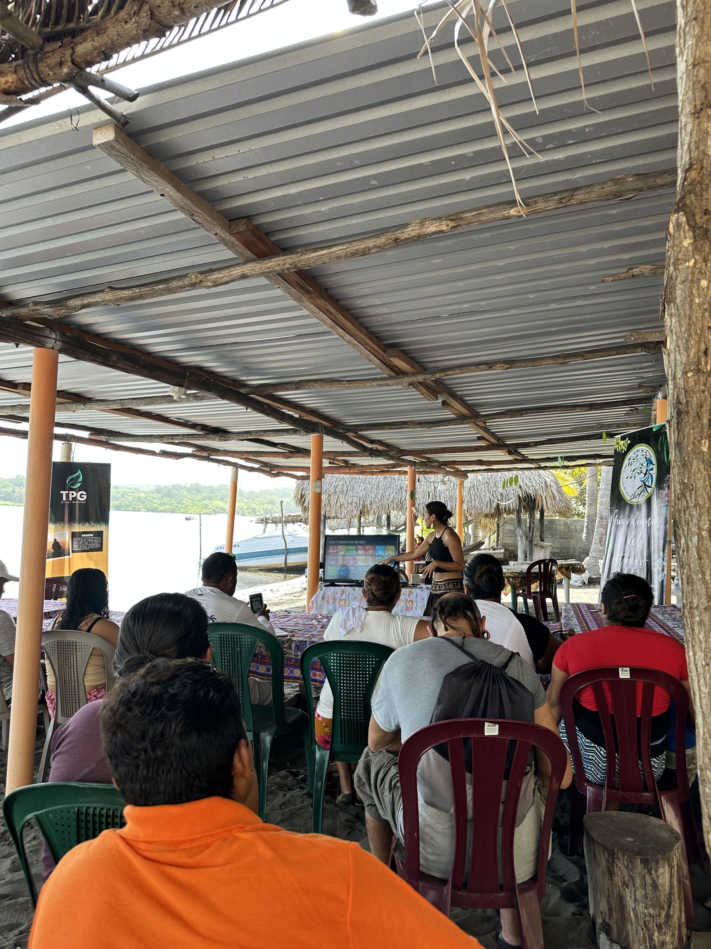 A group of people sit under a metal canopy outdoors, with one person at the front presenting
