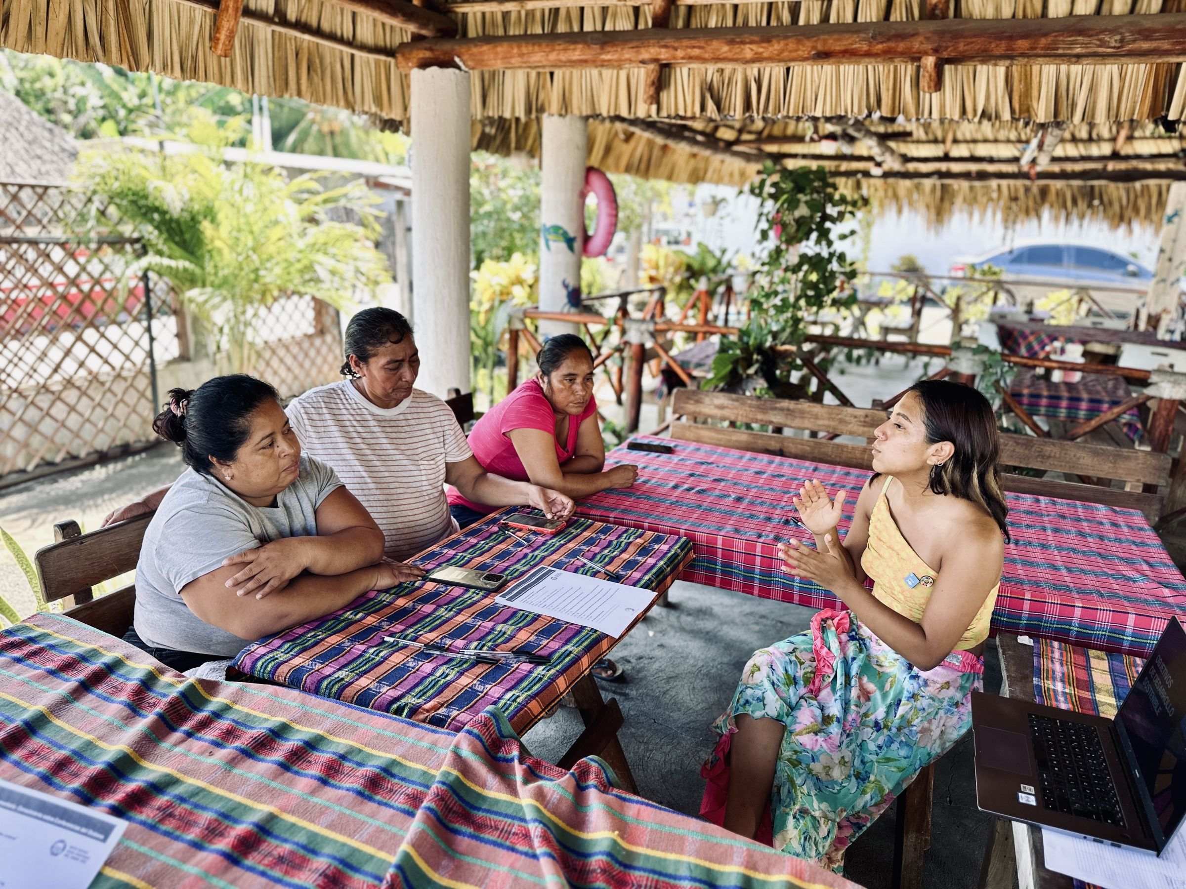 Three people sit at a table in front of another person talking to them next to a laptop, all outdoors under a dried plant and wood canopy