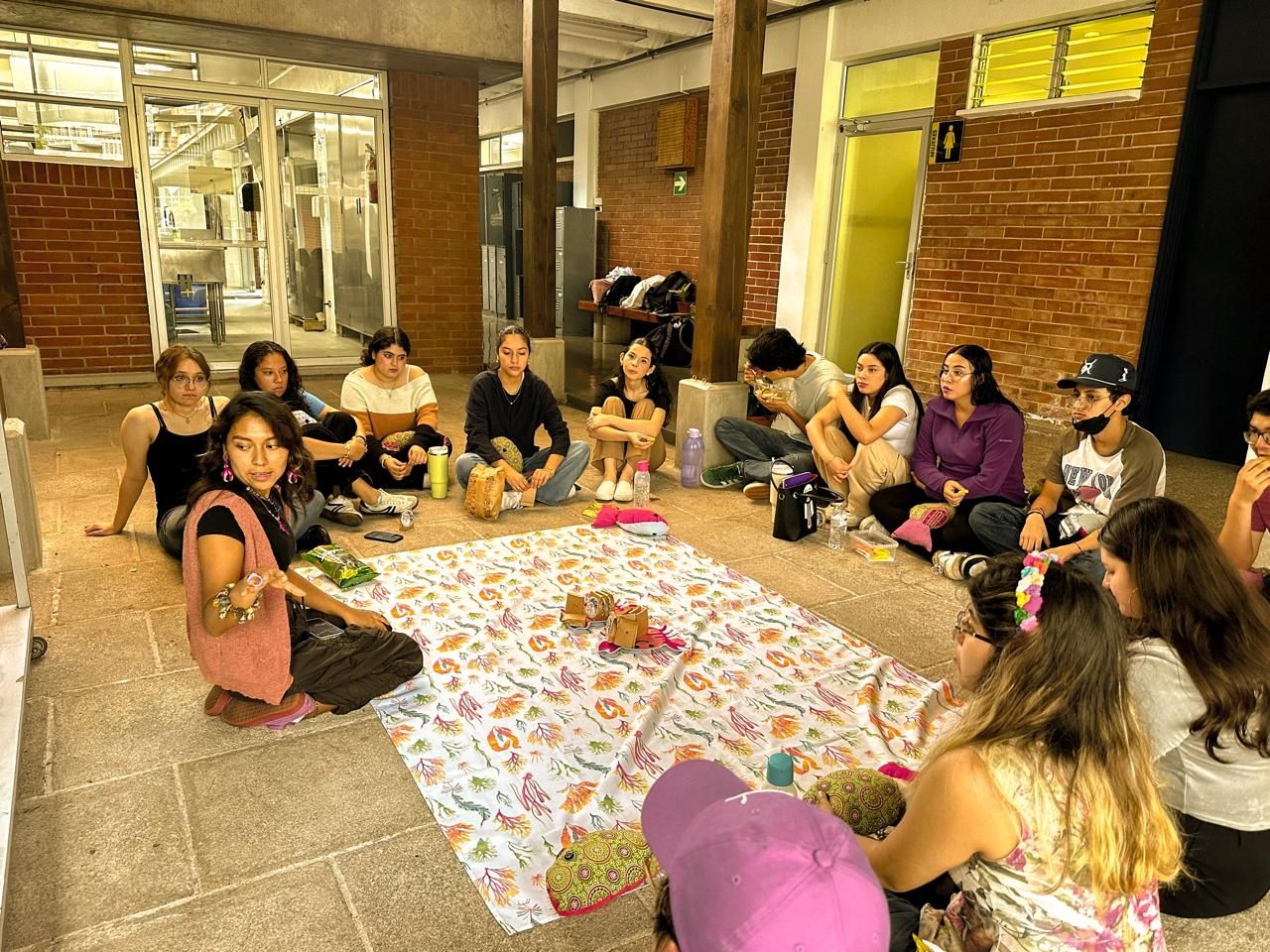A group of students sits around a seaweed-patterned cloth, with one person kneeling right in front of it instructing things