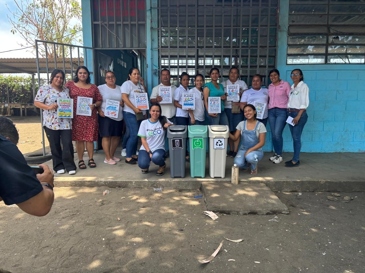 A group of people pose in front of a blue-painted building holding paper; in front of them is a trash can, compost bin, and recycling bin